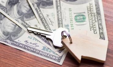 Money with a house key and small wooden house on top