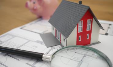 A small red house sitting on blueprints next to a microscope and a piggy bank