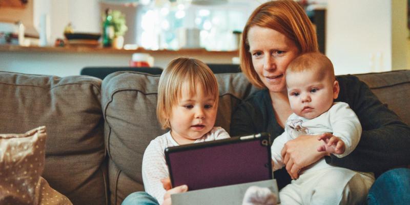 two kids and a woman looking at a tablet on a couch